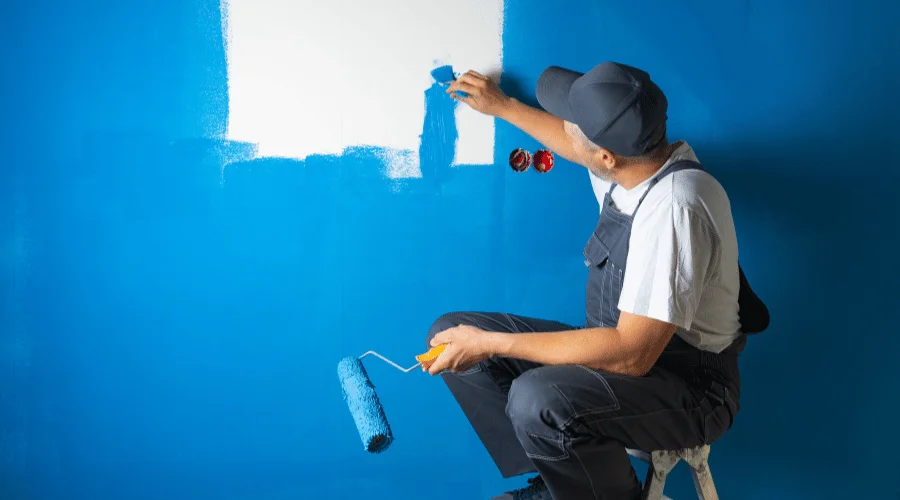 painter using a small brush to paint a big wall while holding a big roller on his other hand